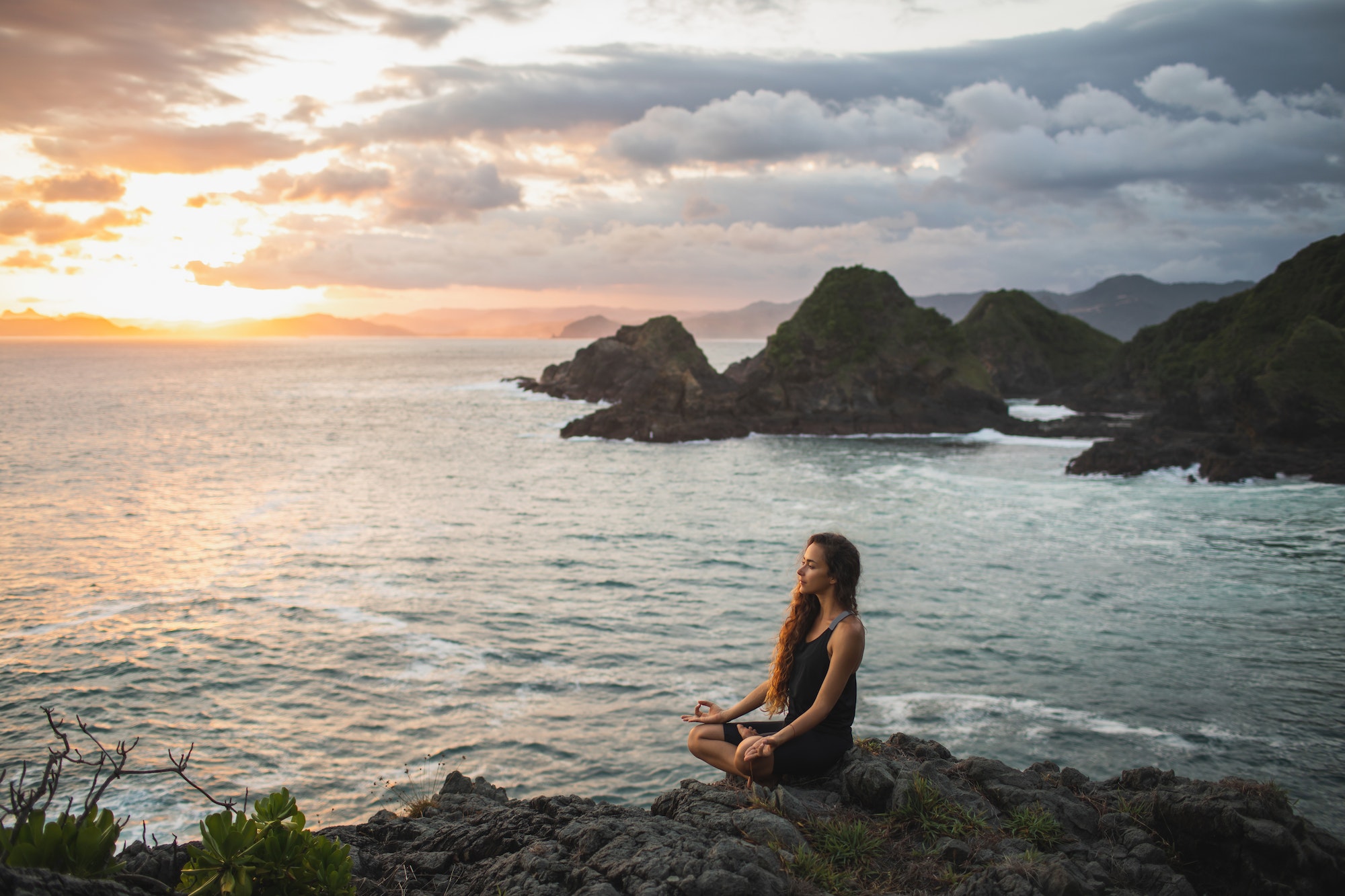 young woman practicing yoga in lotus pose at sunset with beautiful ocean and mountain view