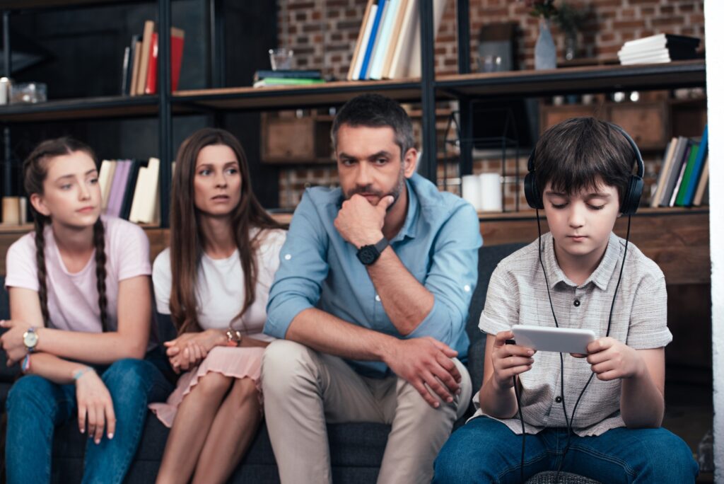 parents and sister looking at addicted boy in headphones using smartphone on family therapy session